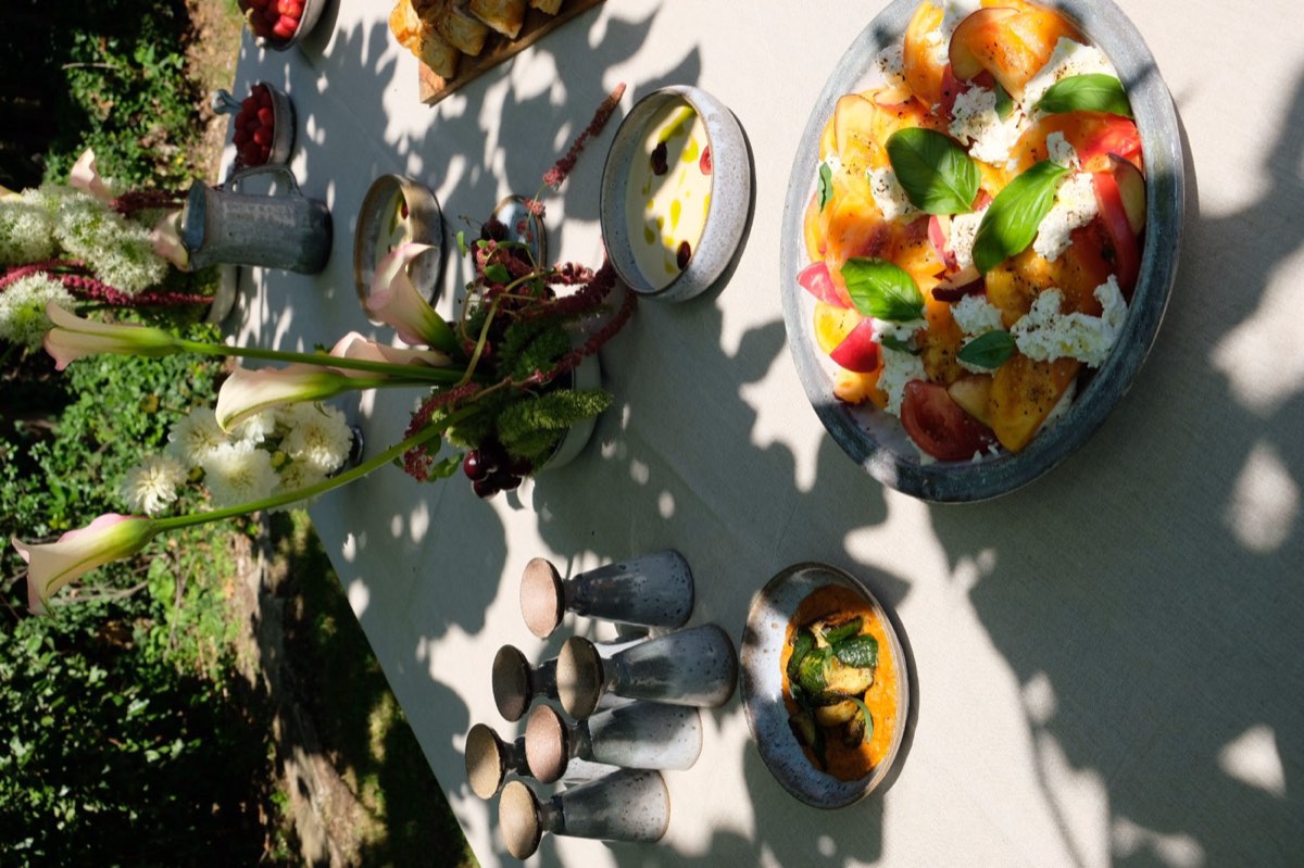Longue table nappée de lin dressée au jardin par tablée sud, avec tomates, fruits d'été, fromage frais et basilic