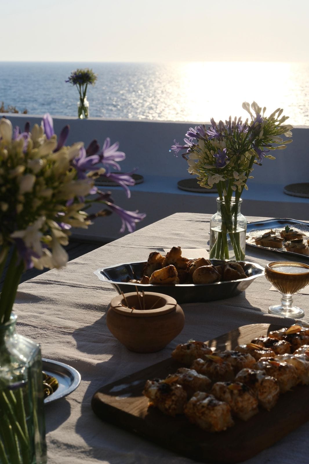 Buffet de tablée sud dressé sur une terrasse face à la mer au coucher du soleil, planches de bouchées et bouquets d’agapanthes